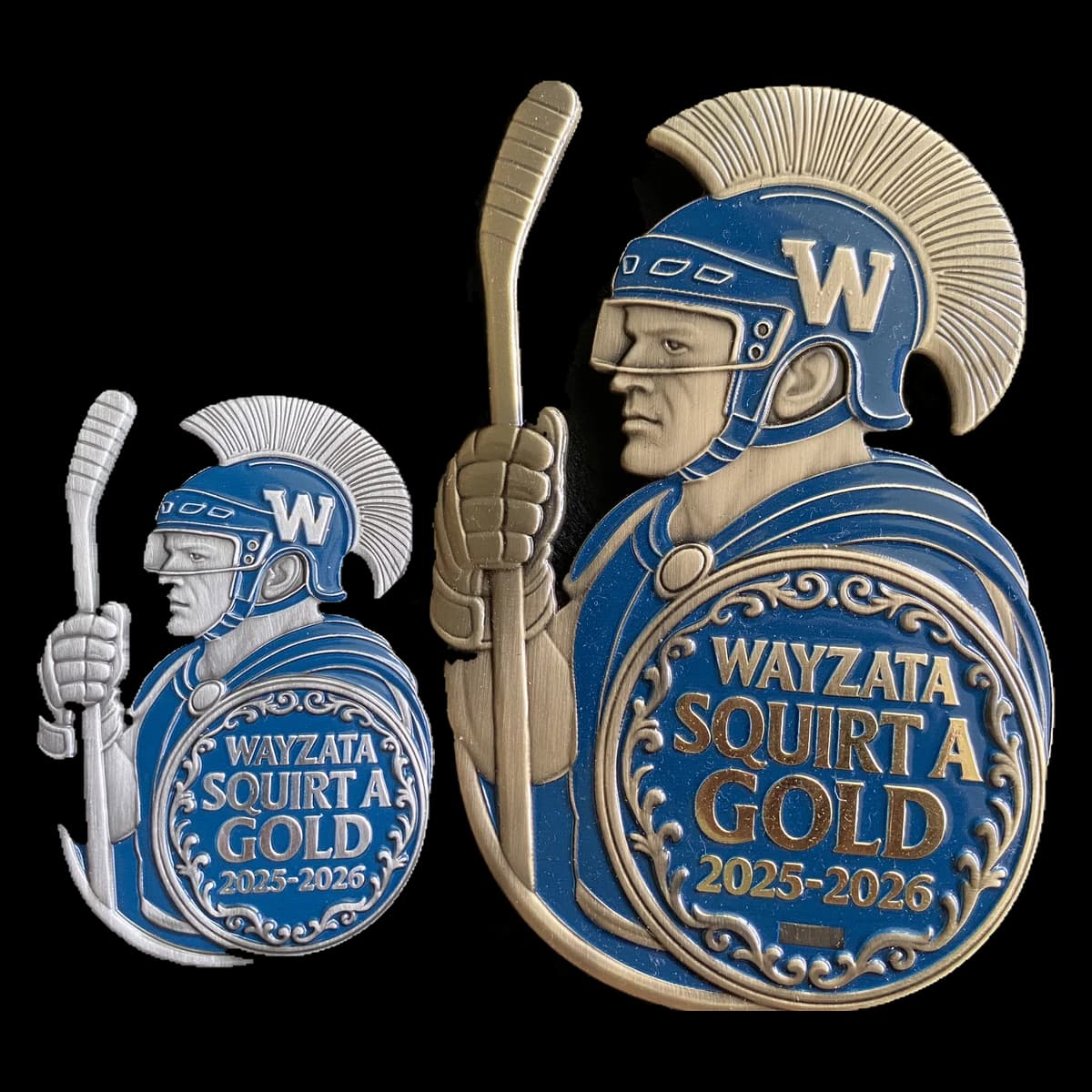 Two Wayzata Squirt A Gold trading pins side by side, a small one in antique silver and a large one in antique gold: a Trojan warrior in a plumed hockey helmet holding a stick, behind a round shield reading Wayzata Squirt A Gold 2025-2026